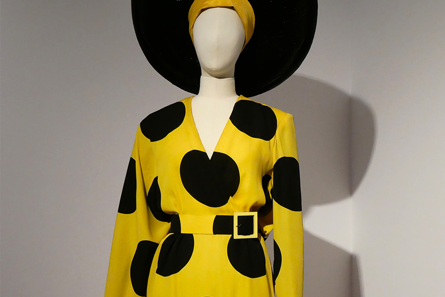 polka dots yellow dress Mannequin wearing a yellow dress with large black polka dots, a matching belt, and a headwrap under a wide-brimmed hat.