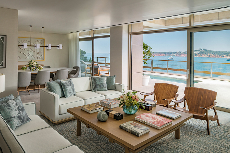 Bright, modern living room with white sofas, wooden chairs, a coffee table with books and flowers, large windows, ocean view, and a connected dining area. Balcony overlooks the water and distant coastline.