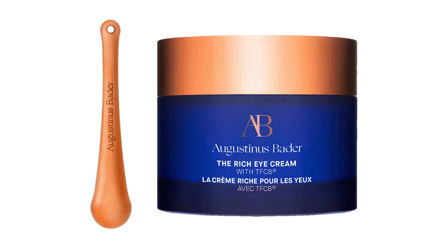 A dark blue jar of Augustinus Bader The Rich Eye Cream with a copper lid, shown next to a matching copper applicator. Both items are displayed against a white background.