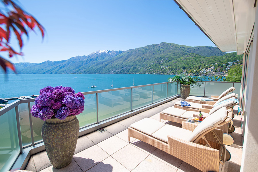 Wicker lounge chairs and potted flowers on a sunny balcony overlook a blue lake with sailboats and lush green mountains under a clear sky.