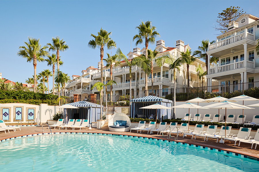 A resort pool with lounge chairs, umbrellas, cabanas, and palm trees, set in front of elegant, white multi-story buildings with balconies on a sunny day.