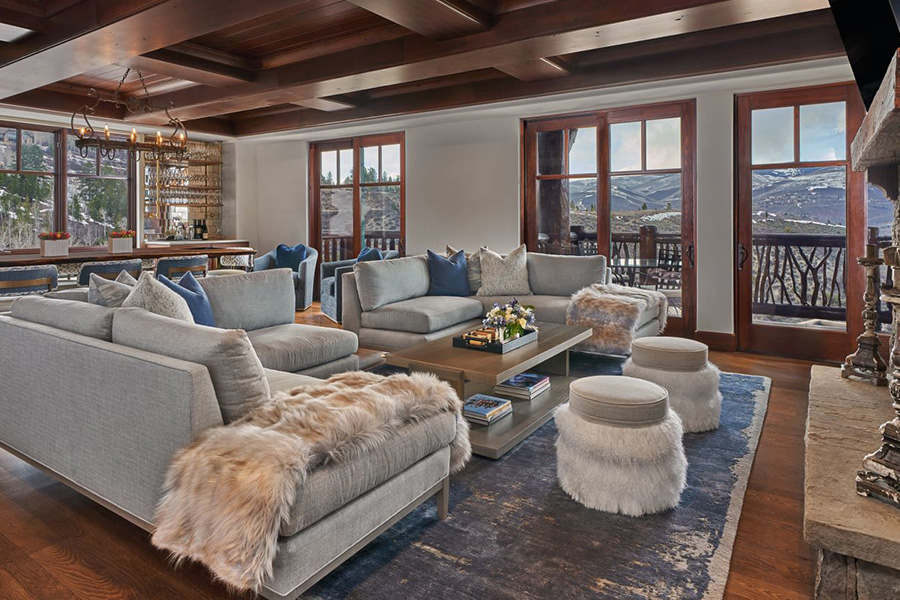 A spacious living room with large windows, grey sectional sofas, fur-covered stools, a blue rug, and a wooden ceiling. The room is bright with natural light and offers scenic mountain views outside.