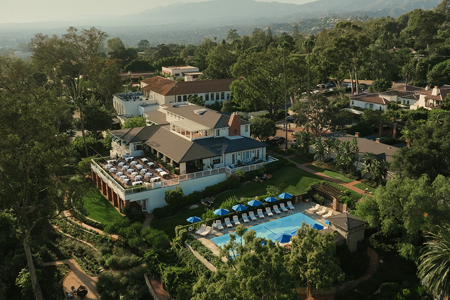 Aerial view of a luxury resort surrounded by lush greenery, featuring a large building with outdoor dining, a swimming pool with blue umbrellas, lounge chairs, and landscaped gardens, set against a backdrop of distant mountains.