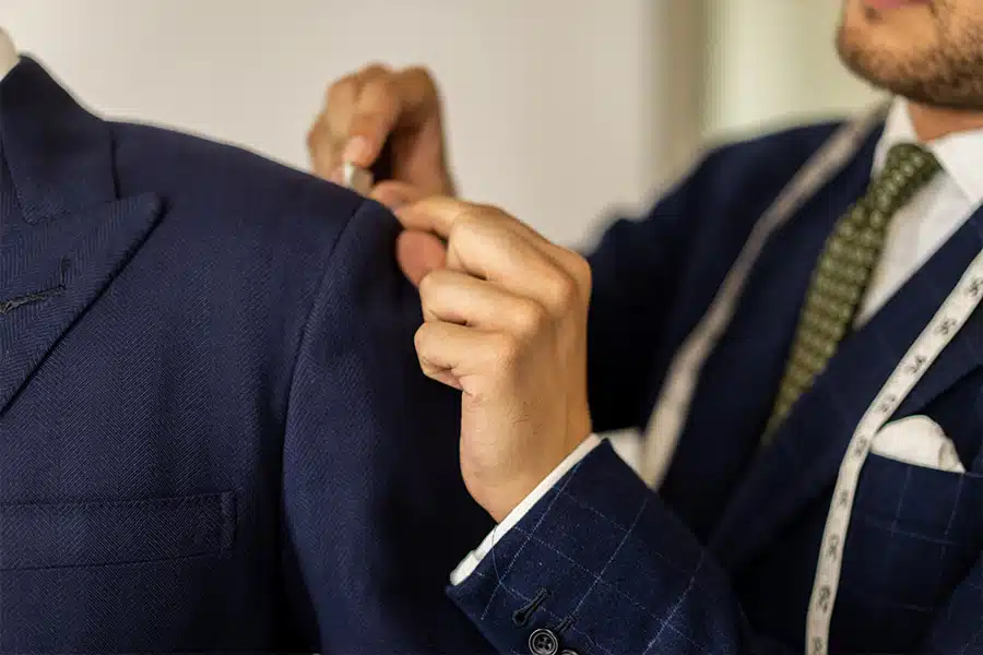 A tailor in a suit measures the shoulder of a navy blue jacket on a mannequin, using a measuring tape draped around his neck.