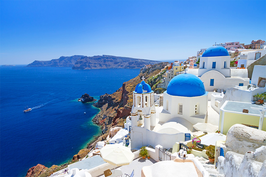 White buildings with blue domes overlook the deep blue sea and rugged cliffs in Santorini, Greece, under a clear sky with a distant boat on the water.