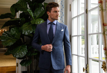 A man in a blue suit with a tie and white pocket square stands indoors near a large window and a tall leafy plant, looking to the side.