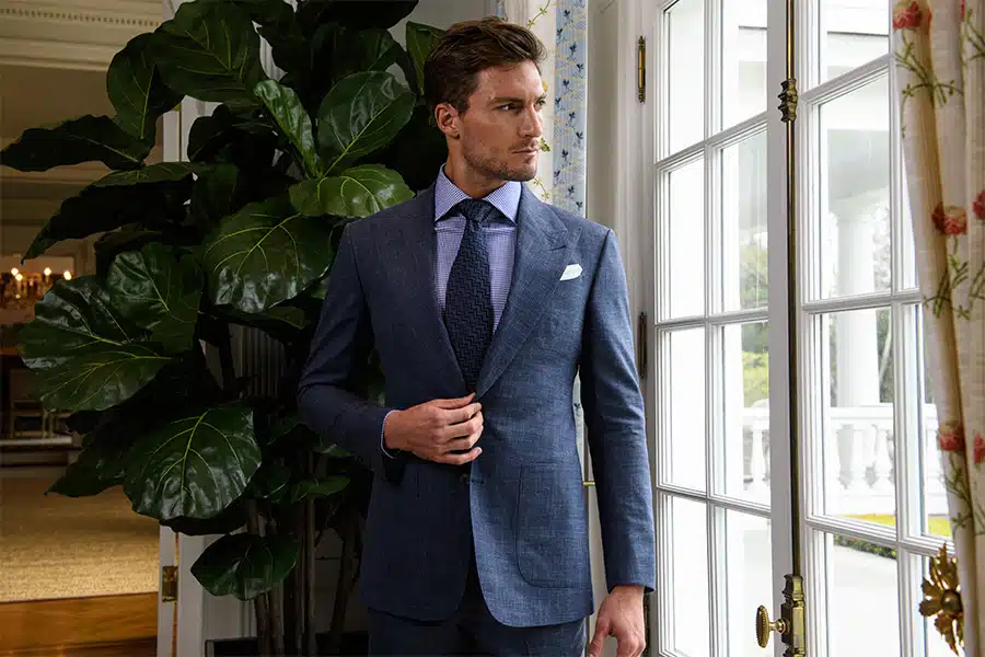 A man in a blue suit with a tie and white pocket square stands indoors near a large window and a tall leafy plant, looking to the side.