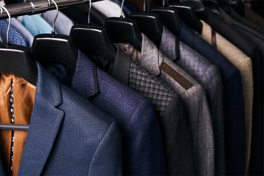 A row of mens suit jackets in various colors and patterns hanging neatly on black hangers in a closet.