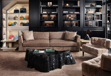 A luxurious living room with a beige sofa, matching armchairs, and a black, irregularly shaped coffee table. In the background are shelves displaying handbags and accessories, all set against dark walls for a sophisticated look.