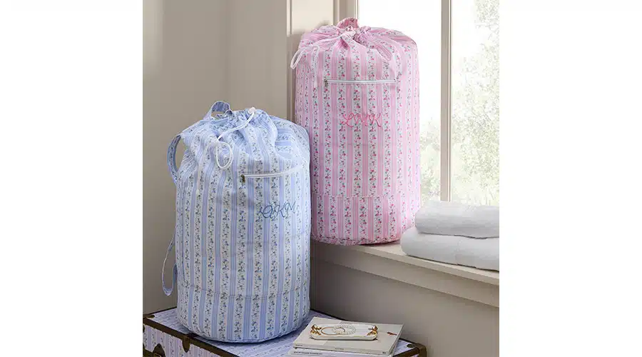 Personalized Laundry Bag Two striped fabric laundry bags, one blue and one pink, sit by a window on a bench next to folded white towels and a closed book. Both bags have drawstring closures and floral patterns.