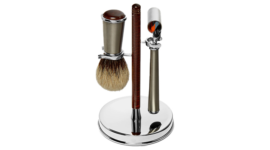 A shaving set with a brush, razor, and stand. The brush has a round bristle head, the razor has a metal handle, and both are held upright on a shiny silver stand with a circular base.