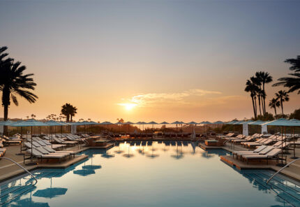 Sunset view over a luxurious outdoor pool surrounded by lounge chairs and umbrellas, with palm trees silhouetted against the sky and the water reflecting the warm light.