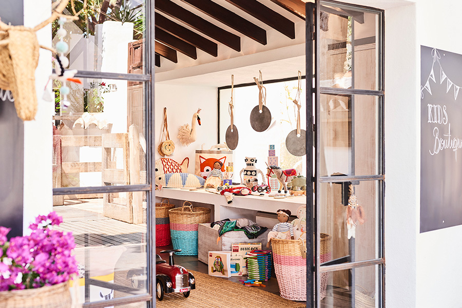Brightly lit kids’ playroom seen through glass doors, featuring soft toys, baskets, books, and musical instruments hanging on the wall. Colorful, cozy, and inviting space with flowers in the foreground.