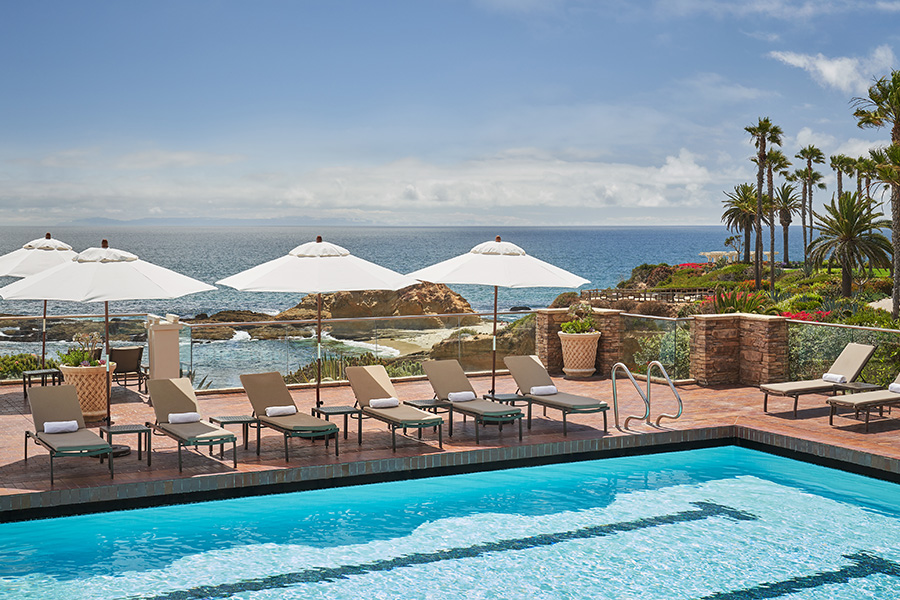 Lounge chairs and white umbrellas line a poolside terrace overlooking rocky shores and the ocean. Palm trees and lush landscaping enhance the coastal, resort-like atmosphere on a sunny day.