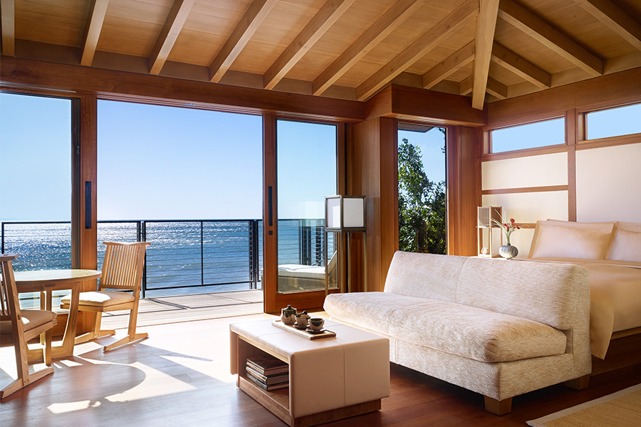 A bright, modern bedroom with wooden beams, a cream sofa, a bed, a small dining table, and large glass doors opening to a balcony overlooking the ocean. Sunlight fills the room, creating a warm, inviting atmosphere.