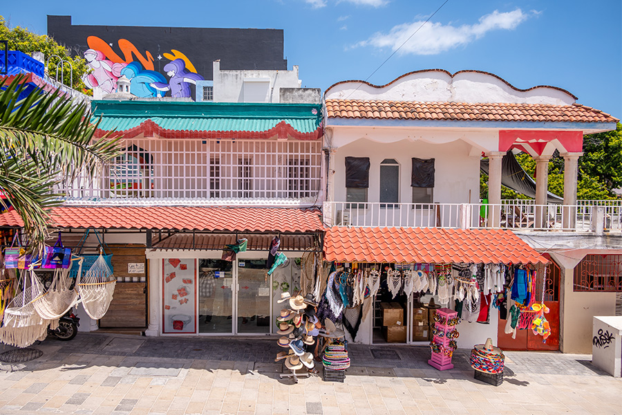 Colorful streetfront shops with red tile roofs display hats, hammocks, bags, and souvenirs. The upper level has white railings and artistic murals, with a bright blue sky in the background.