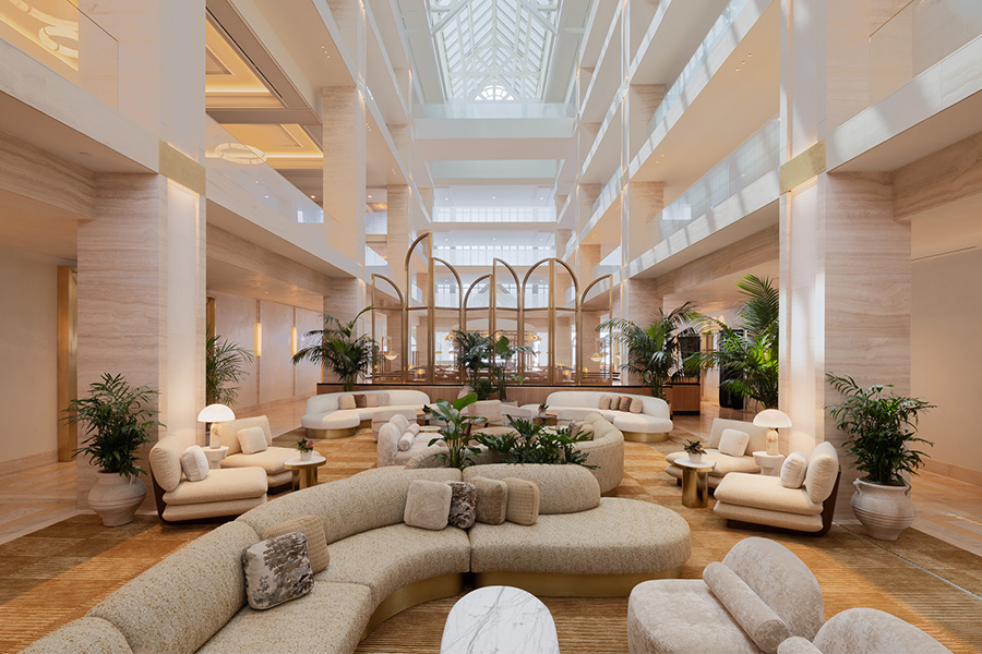 Spacious, modern hotel lobby with cream-colored curved sofas, round tables, indoor plants, soft lighting, and high atrium ceiling with large skylight and balconies on multiple floors.