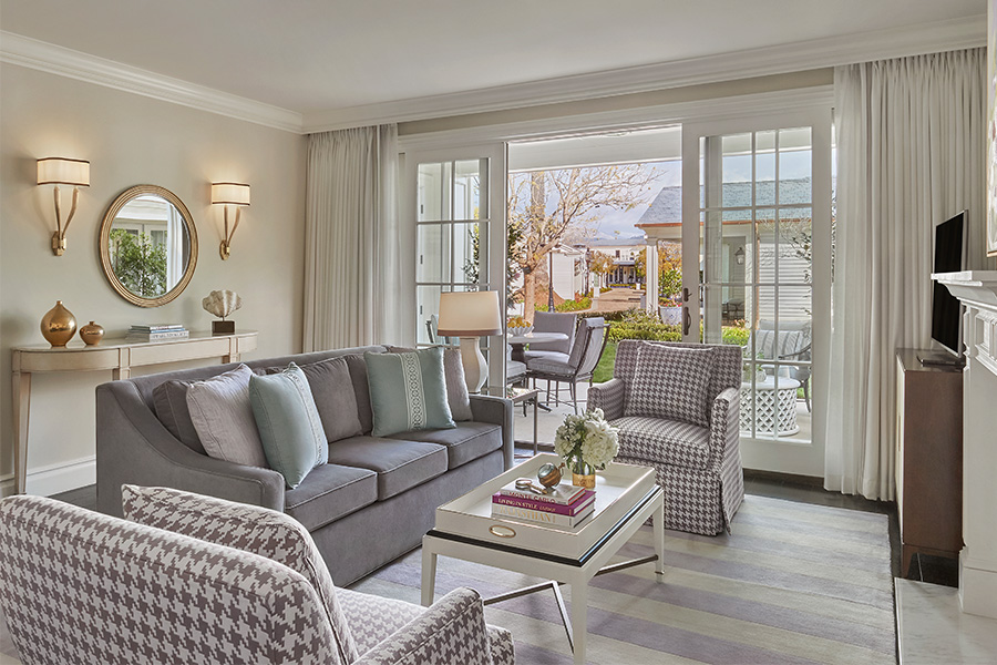 Bright living room with gray sofa, patterned armchairs, a glass coffee table, and decorative items. Large windows and glass doors open to a sunny patio, letting in natural light. Neutral tones and elegant decor create a cozy atmosphere.