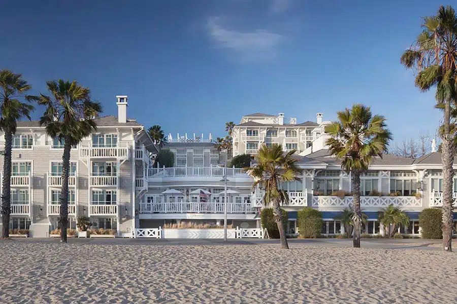 Elegant beachfront hotel with white wooden railings, palm trees, and sandy beach in the foreground under a clear blue sky.