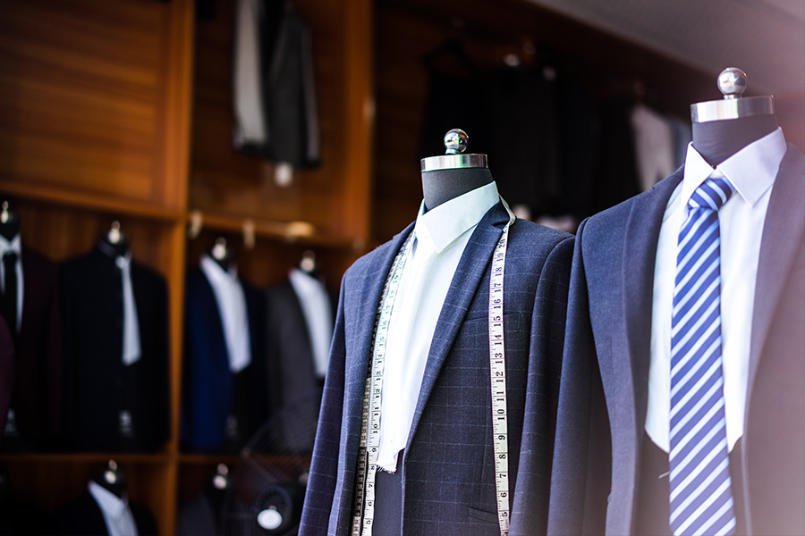 Three mannequins in suits are displayed in a tailor shop, with measuring tape draped around one. Blurred shelves with more suits and ties are visible in the background.