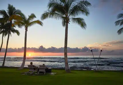 Two people sit in lounge chairs on a grassy area by the ocean, watching the sunset. Palm trees frame the view, and two tiki torches stand near the shoreline. The sky is colorful with clouds and a glowing sun.