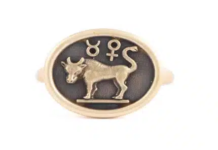 An oval gold ring featuring a raised bull, the Taurus zodiac symbol, with Taurus and Venus symbols above it on a dark background.