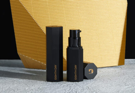 Two black bottles of Eighth Day skincare with gold text stand on a textured gray surface, one open with a pump exposed, in front of a large gold, geometric background.