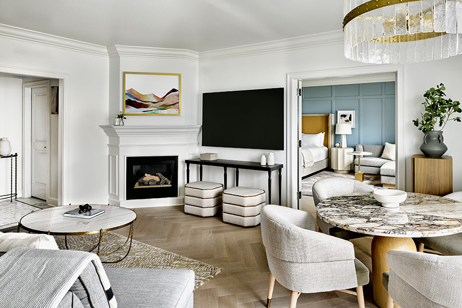 Modern living room with light neutral tones, a fireplace with abstract art above, a large flat-screen TV, plush seating, a round marble dining table, and an adjoining bedroom visible through open double doors.