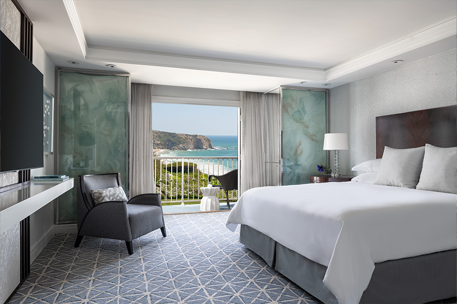 A modern hotel room with a large bed, armchair, TV, and lamps. Sliding doors open to a balcony with a view of the ocean, cliffs, and blue sky. The room has light colors and geometric-patterned carpet.