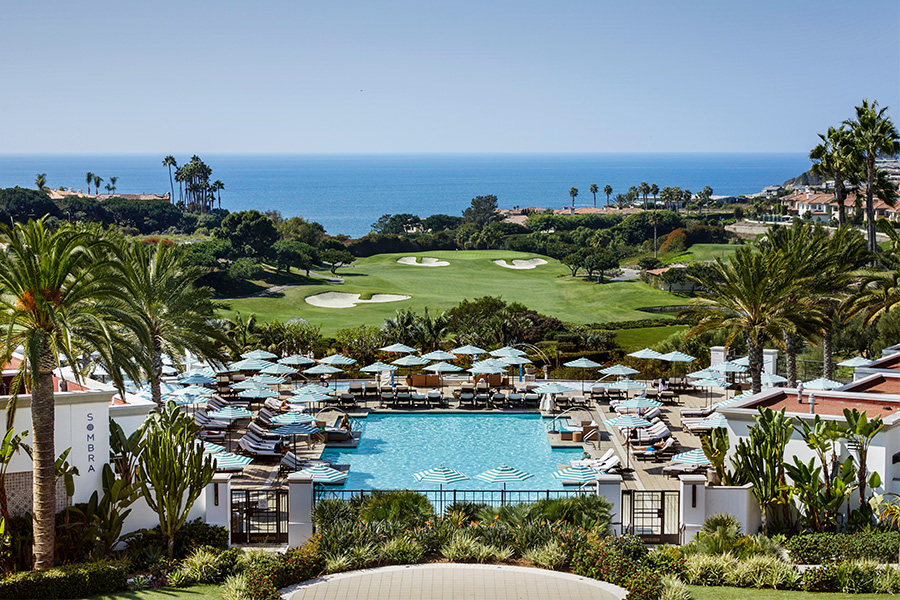 A luxurious resort with a large swimming pool surrounded by lounge chairs and umbrellas, overlooking a lush golf course and the ocean in the distance under a clear blue sky.