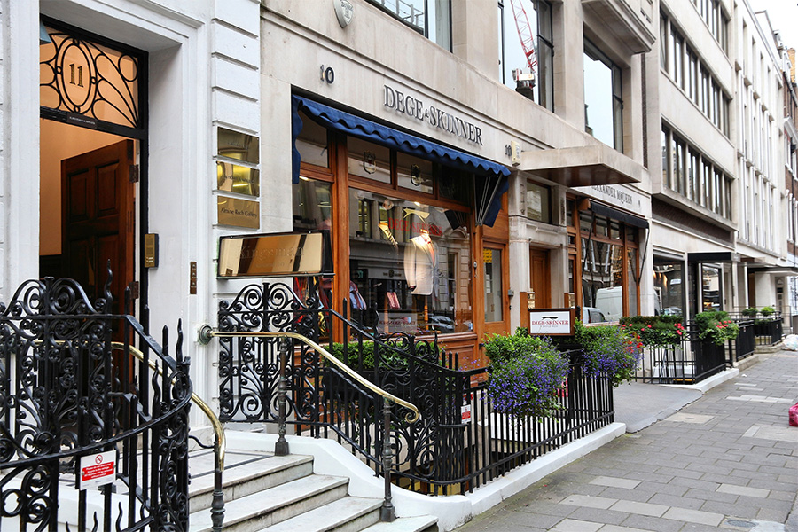 A view of a classic storefront for Dege & Skinner at 10 Savile Row, London. The shop features wooden-framed windows, a blue awning, railings, steps, and flower boxes along the entrance.