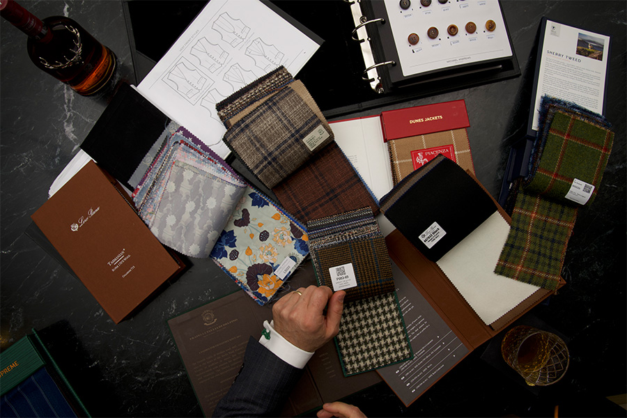 A person’s hand examines various fabric swatches and pattern books on a dark table, surrounded by a glass of whiskey, a bottle, paperwork, and open binders with fabric information and diagrams.