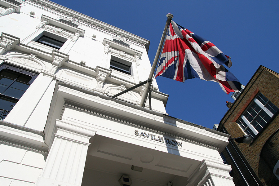 A Union Jack flag flies above the white entrance of a building with Savile Row written above the door, set against a clear blue sky.