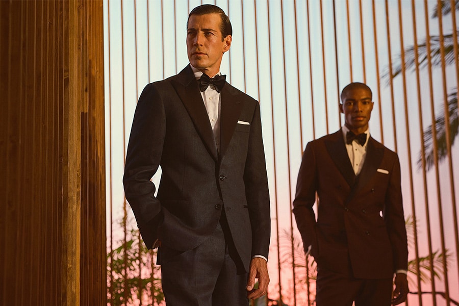 Two men wearing tuxedos stand in front of vertical wooden slats at sunset; one wears a black tuxedo and bow tie, the other a brown tuxedo, both with white shirts and pocket squares.