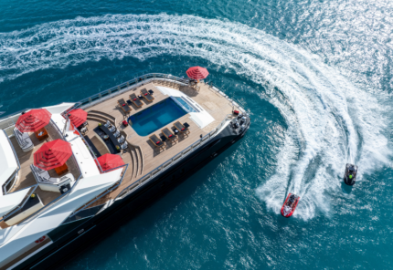 Aerial view of a luxury yacht with a pool and red umbrellas on deck, anchored in clear blue water. Two jet skis create white trails as they speed past the yacht.
