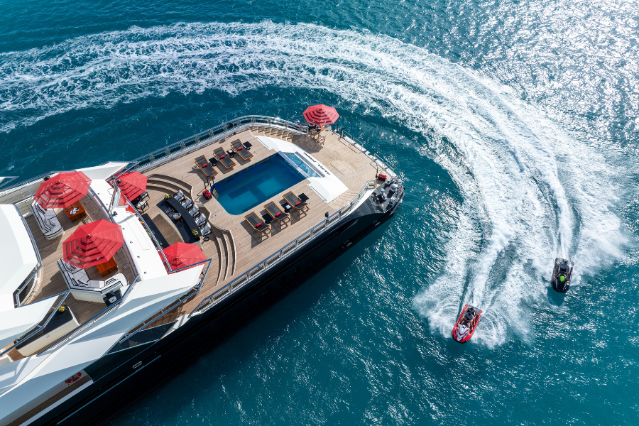 Aerial view of a luxury yacht with a pool and red umbrellas on deck, anchored in clear blue water. Two jet skis create white trails as they speed past the yacht.
