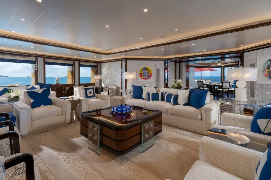 Luxurious yacht interior with cream-colored sofas, blue accent pillows, a glossy wood coffee table, large windows showing ocean views, and elegant decor including lamps, flowers, and modern art on the walls.