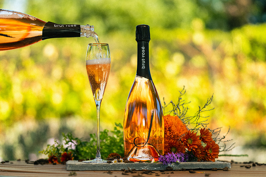A bottle of brut rosé is poured into a champagne flute on an outdoor table, next to another full bottle and vibrant orange flowers, with a blurred green background.