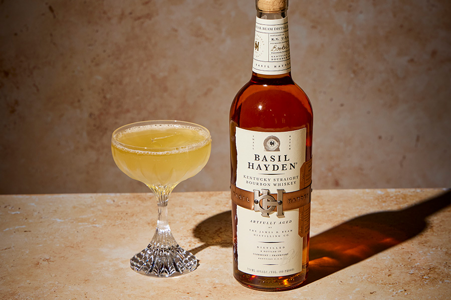 A bottle of Basil Hayden Kentucky Straight Bourbon Whiskey stands next to a coupe glass filled with a light yellow cocktail on a beige surface with a matching beige background.