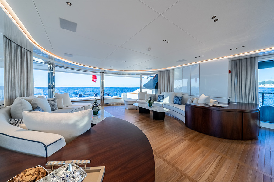 Luxurious yacht interior with curved white sofas, wooden flooring, and large windows offering panoramic ocean views. Elegant decor, natural light, and blue sea create a spacious, relaxing atmosphere.