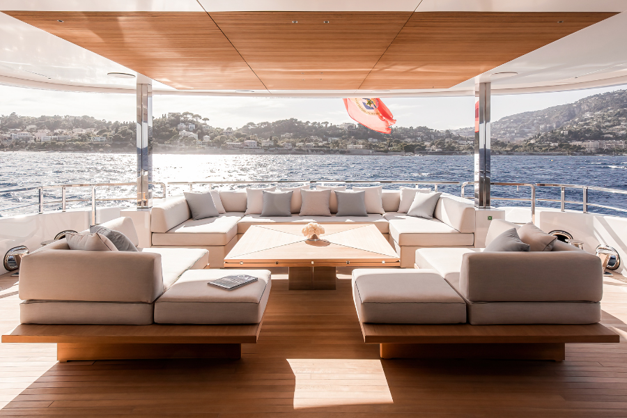 A luxurious yacht deck with modern, beige cushioned seating arranged in a U-shape, a wooden table in the center, and a scenic ocean view with a hilly coastline and a red flag at the back.