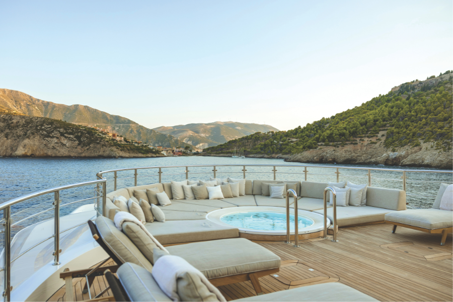 Luxurious yacht deck with cushioned seating and a round hot tub, surrounded by clear blue water and scenic green hills under a bright sky.