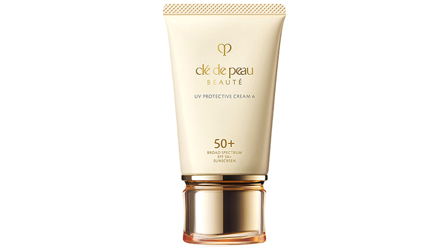 A tube of Clé de Peau Beauté UV Protective Cream with SPF 50+ is shown. The gold and white packaging features the brand name, product name, and sun protection details in gold text.