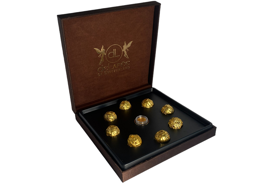 An open brown box with gold lettering on the inside lid displays eight gold-wrapped chocolates arranged in a circle around a small, clear container in the center.