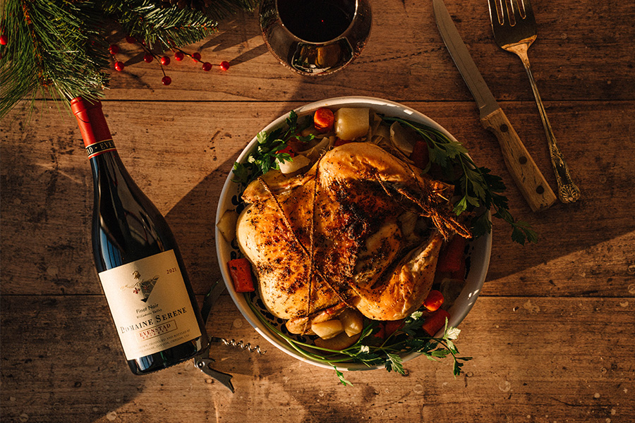 A roasted chicken garnished with herbs and vegetables sits on a wooden table beside a bottle of red wine, a glass of wine, a carving knife, and festive holiday greenery.