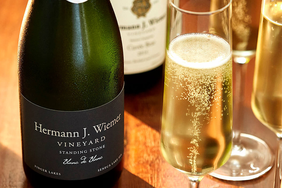A bottle of Hermann J. Wiemer Vineyard sparkling wine is displayed beside filled champagne flutes on a wooden surface, with bubbles rising in the glasses and sunlight highlighting the scene.