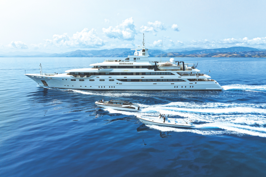 A large luxury yacht cruises on calm blue water, followed by two smaller motorboats creating white wakes. Distant mountains and a partly cloudy sky are visible in the background.