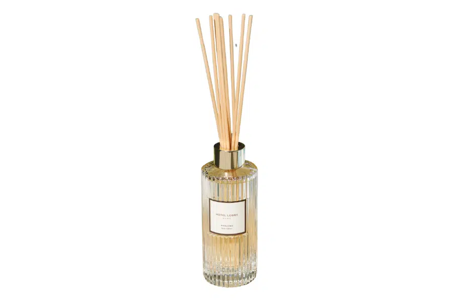 A clear, ribbed glass diffuser bottle with light brown fragrance sticks and a gold cap, filled with light yellow liquid and a white label on the front.