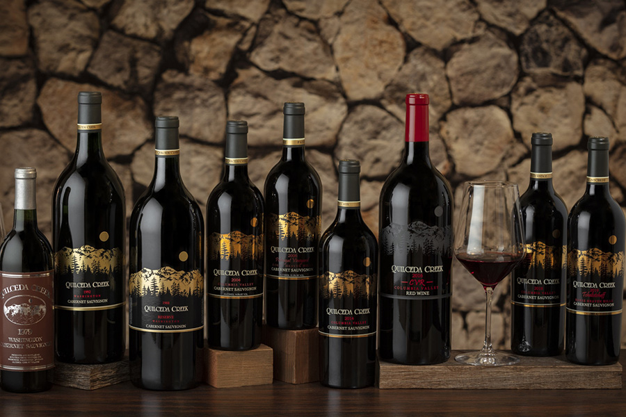 A group of Quixote wines in dark bottles with gold and red labels are arranged on a wooden table in front of a stone wall, alongside a filled wine glass.