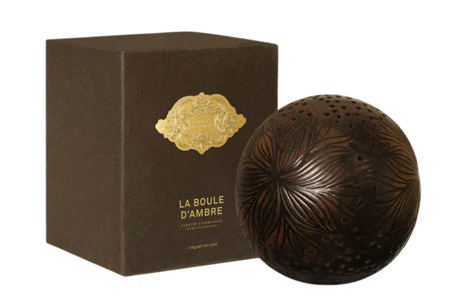 A decorative brown sphere with floral carvings sits beside a dark brown box labeled “La Boule d’Ambre” in gold, with a gold emblem above the text.